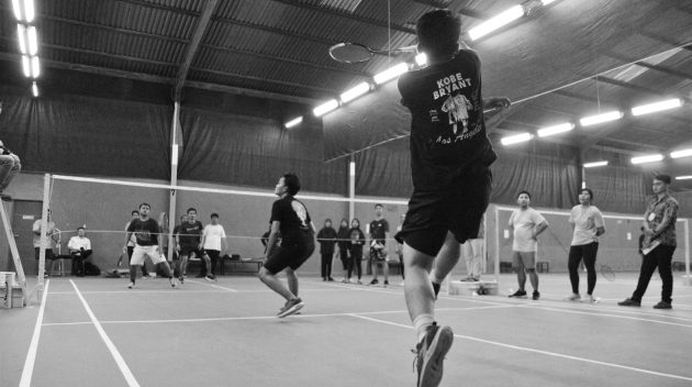 Black and white image capturing intense indoor badminton match with players and spectators.