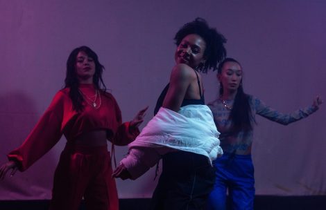 Three female dancers perform dynamically against a purple-lit background, showcasing vibrant choreography and diversity.