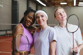 Three women smiling, holding badminton rackets in an indoor court, showcasing active lifestyle.